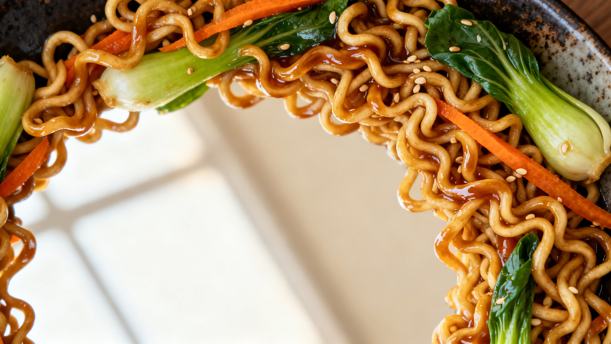 Overhead view of ramen noodle stir fry with glossy soy-sesame sauce, green bok choy, and orange carrot ribbons in a dark ceramic plate.