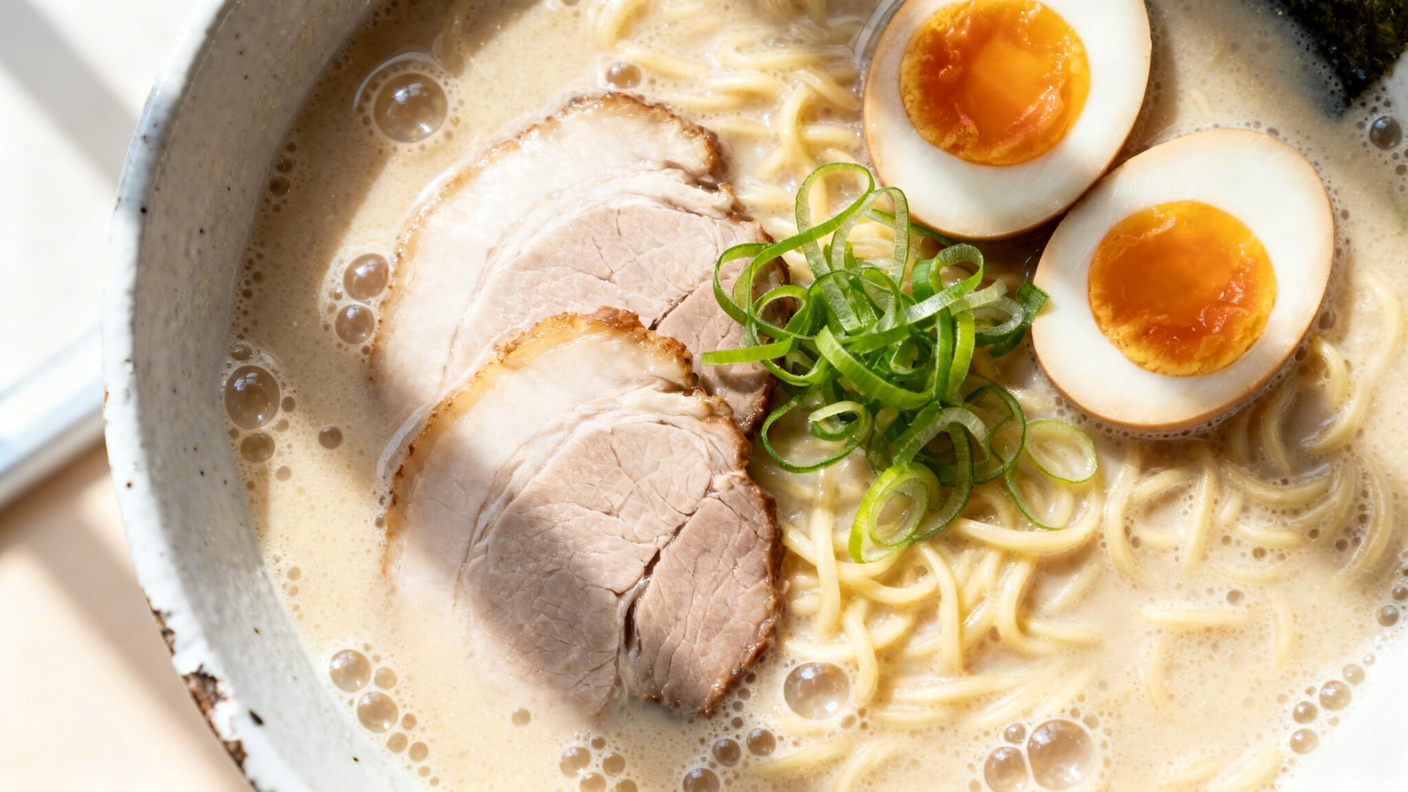Overhead view of tonkotsu ramen in a white ceramic bowl with milky broth, chashu pork slices, a halved soft-boiled egg, and green onions.