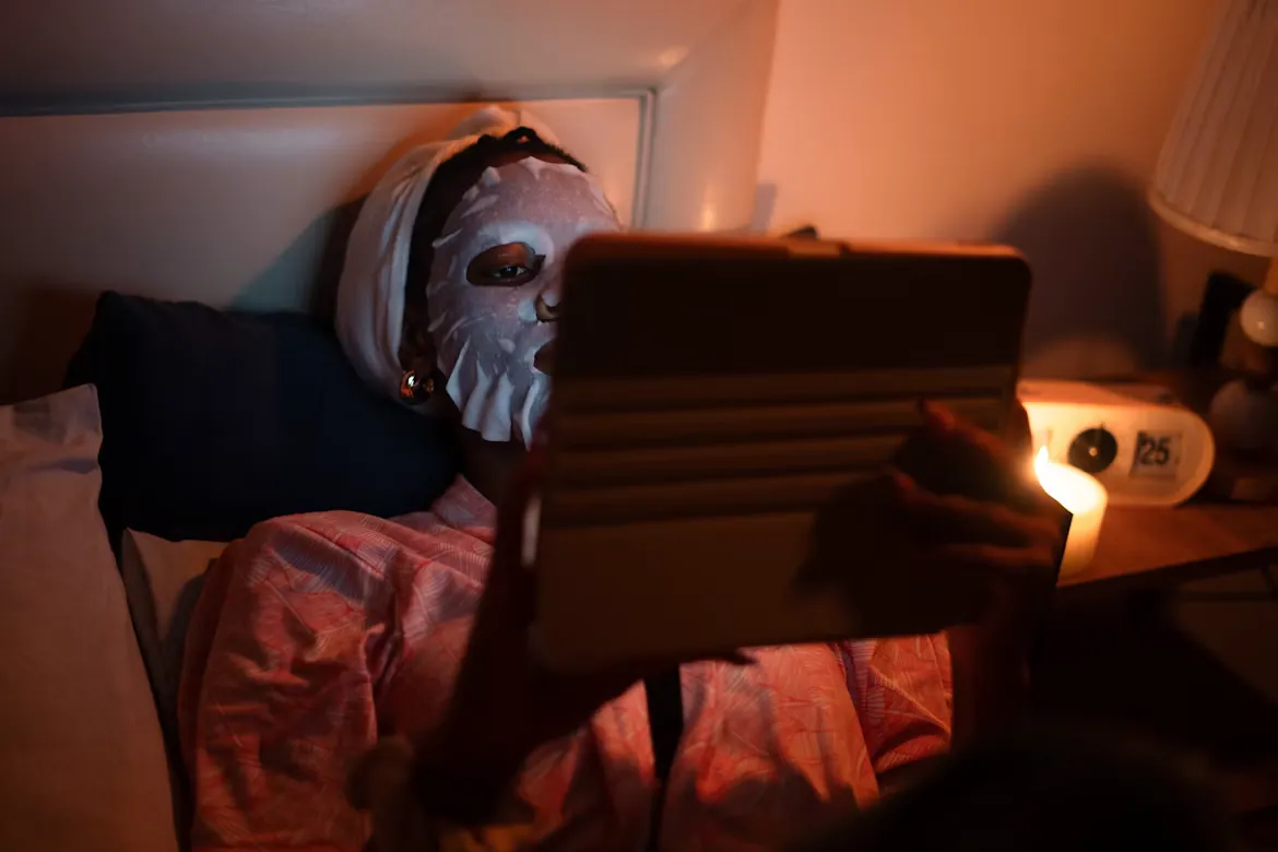 Woman in a sheet face mask relaxing in bed with a tablet by candlelight